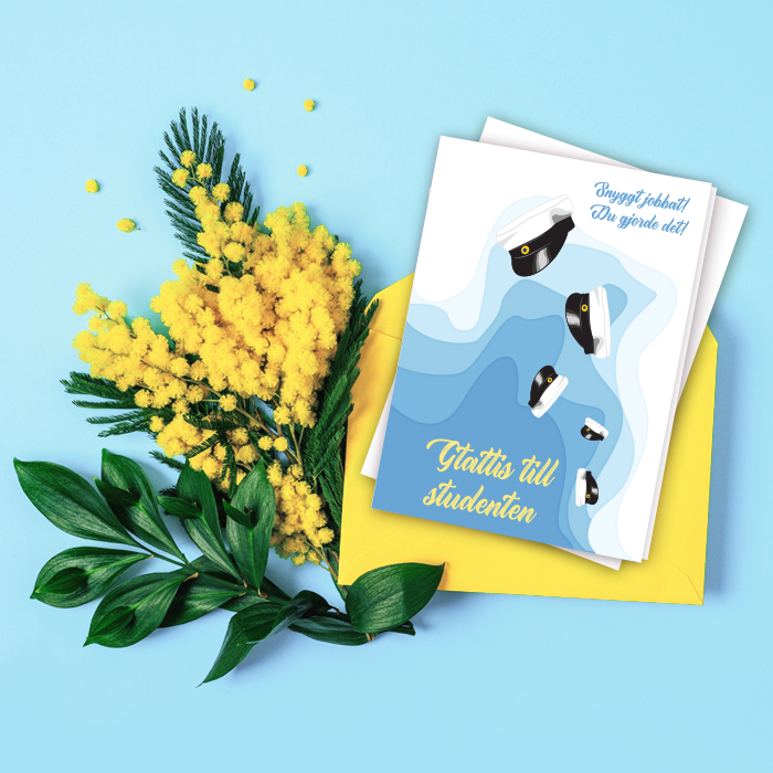 Ett blått och vitt kort med flygande studentmössor, liggandes på en blå bakgrund med gula blommor