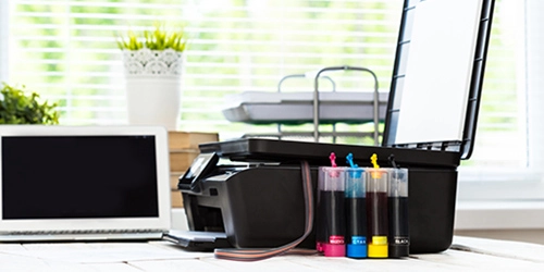 Une imprimante avec le couvercle ouvert est posée sur une table à côté d’un ordinateur portable. Devant l’imprimante se trouvent des réservoirs d’encre de plusieurs couleurs. Une imprimante avec le couvercle ouvert est posée sur une table à côté d’un ordinateur portable. Devant l’imprimante se trouvent des réservoirs d’encre de plusieurs couleurs.