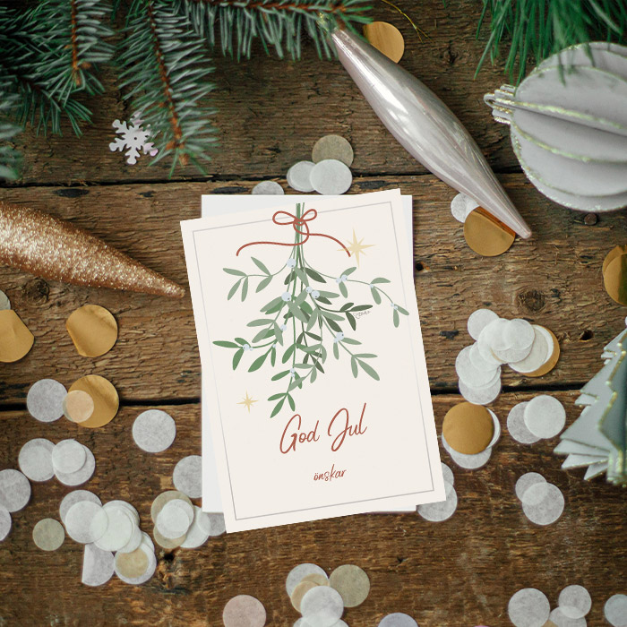 Ett kort med mistel och texten god jul liggandes på ett golv med konfetti och julgranskulor runt omkring