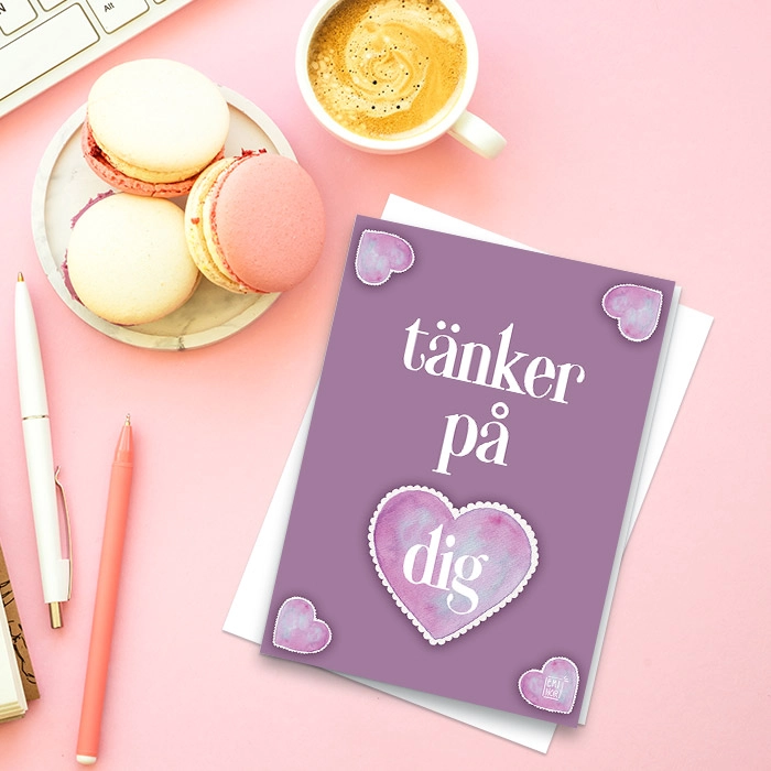 Ett lila kort med hjärtan som har texten jag tänker på dig, liggandes på ett skrivbord med fika