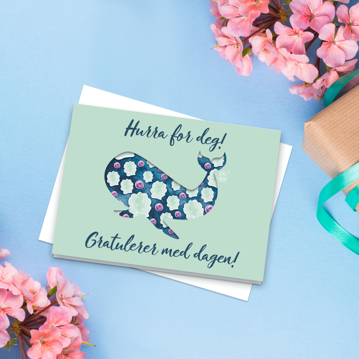 Gratulasjonskort med en hval på blå bakgrunn, et bord med blomster og en gave.