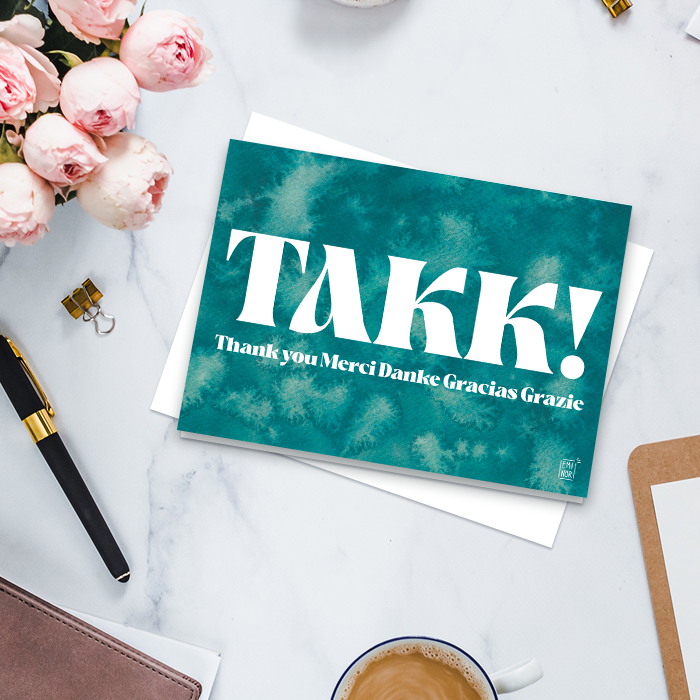 Takkekort med teksten takk på forskjellige språk, med en turkis akvarellbakgrunn, liggende på et skrivebord.