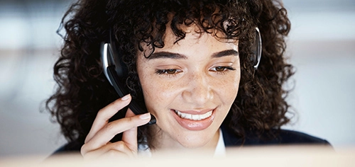 Une employée du service client souriante, portant un casque, parle avec un client devant un ordinateur. Une employée du service client souriante, portant un casque, parle avec un client devant un ordinateur.