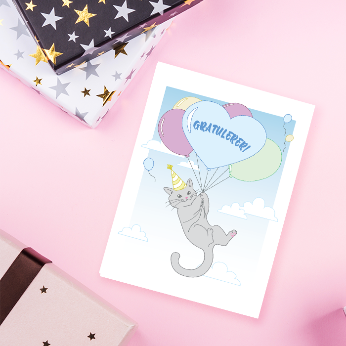 Gratulasjonskort med en katt som holder ballonger med teksten Gratulerer, liggende på et bord med gaver.
