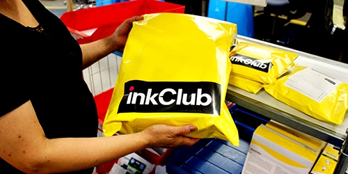 Une personne tient un colis jaune portant le logo inkClub, sur une table d’emballage avec d’autres colis similaires à l’arrière-plan. Une personne tient un colis jaune portant le logo inkClub, sur une table d’emballage avec d’autres colis similaires à l’arrière-plan.